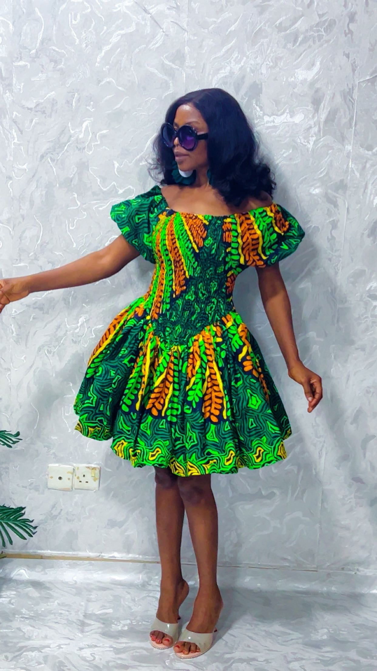 Woman wearing a colorful dress with a green and yellow pattern against a textured white wall.
