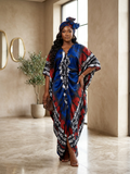 A woman models a vibrant blue, red, and black patterned kaftan dress with matching headwrap in a modern, neutral-toned indoor setting.